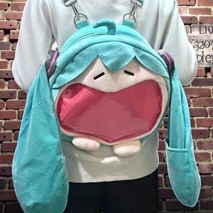May include: A teal plush backpack shaped like a cartoon character's face with large, open mouth. The backpack has long teal pigtails, pink accents, and a clear pocket in the mouth area. It has adjustable straps and metal hardware.