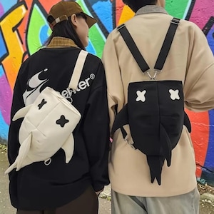 May include: Two backpacks shaped like sharks, one in white and one in black. Each bag features a dorsal fin, side fins, and two white or black cross-shaped eyes. The white backpack is worn by a person in a black shirt, and the black backpack is worn by a person in a beige shirt, both with adjustable straps.