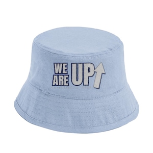 Może przedstawiać: Jasnoniebieski kapelusz typu bucket z napisem "WE ARE UP" w kolorze granatowym i białym. Kapelusz ma zaokrągloną koronę i opadający rondo. Kapelusz jest wykonany z miękkiej, teksturowanej tkaniny.