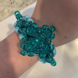 Op de afbeelding: Een kralenarmband in tinten turquoise en lichtblauw. De armband heeft een cluster van grotere, transparante kralen in het midden, omringd door kleinere, ondoorzichtige kralen. Het ontwerp is een mix van ronde en vierkante kralen, wat een textuur geeft.