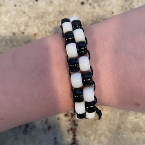 Può includere: Un braccialetto di perline bianche e nere con un motivo a scacchiera. Il braccialetto è composto da piccole perline cilindriche infilate su un cordino nero. Le perline si alternano tra bianco e nero, creando un design grafico audace.