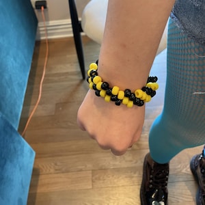 Puede incluir: Un brazalete hecho a mano con cuentas amarillas y negras. El brazalete se lleva en la muñeca, destacando el contraste de colores y el patrón repetitivo de las cuentas. Un accesorio sencillo pero llamativo.