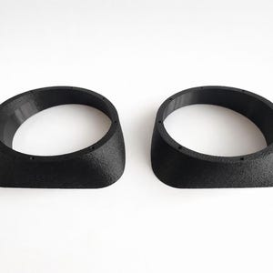 May include: Two black, oval-shaped objects with a textured surface. Each object has a circular opening and a slightly angled side. The objects are placed side by side against a white background.