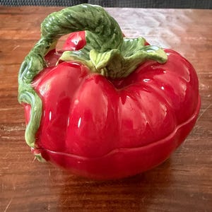 May include: A ceramic tomato-shaped container with a glossy red finish. The lid features a green stem and leaves, adding a naturalistic touch. This decorative piece is likely used for serving or storing food.