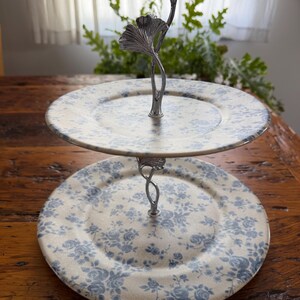 May include: Two-tiered serving stand with floral patterned plates. The plates have a cream background with blue floral designs. The stand is made of silver metal and features a decorative ginkgo leaf design at the top.