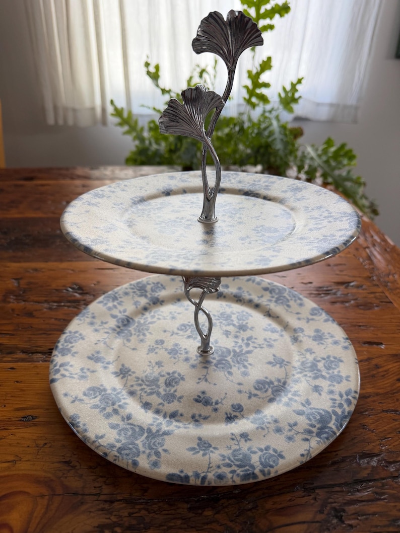 May include: Two-tiered serving stand with floral patterned plates. The plates have a cream background with blue floral designs. The stand is made of silver metal and features a decorative ginkgo leaf design at the top.