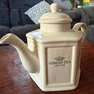 May include: Cream-colored ceramic teapot with a square body and a curved spout. The teapot has a lid with a small knob and a handle. The front of the teapot features the text "AHMAD TEA LONDON" in black.