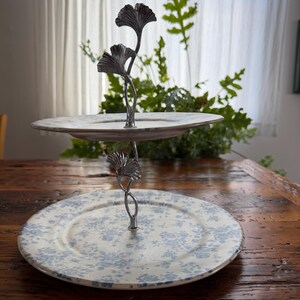 May include: Two-tiered serving stand with a metal ginkgo leaf design. The plates have a cream background with blue floral patterns. The stand is on a wooden table, with greenery in the background.