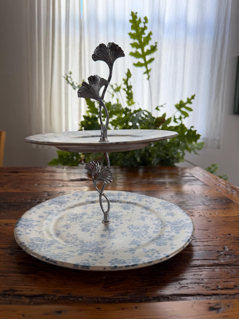 May include: Two-tiered serving stand with a metal ginkgo leaf design. The plates have a cream background with blue floral patterns. The stand is on a wooden table, with greenery in the background.