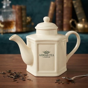 May include: A cream-colored hexagonal ceramic teapot with a matching lid, featuring the black text AHMAD TEA LONDON and a small crown logo on the front. The teapot has a curved handle and a spout, with loose tea leaves scattered on the surface.