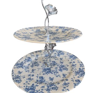 May include: Two-tiered serving tray with a silver-colored handle featuring a ginkgo leaf design. The round plates have a cream background with a blue floral pattern. Ideal for serving desserts or appetizers.