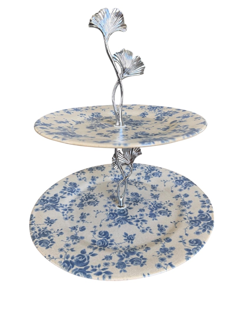 May include: Two-tiered serving tray with a silver-colored handle featuring a ginkgo leaf design. The round plates have a cream background with a blue floral pattern. Ideal for serving desserts or appetizers.