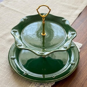May include: A two-tiered serving stand made of glossy dark green ceramic. The plates feature scalloped edges with small raised circular details along the rims. A central gold-toned metal handle connects the two tiers at the top.