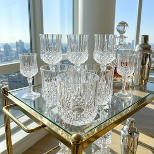May include: A collection of clear cut-glass barware including stemmed wine glasses, short tumblers, and a tall decanter with a stopper, arranged alongside a metallic cocktail shaker on a gold-toned metal bar cart with glass shelves.