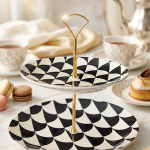 May include: A two-tiered serving stand featuring two round plates with a black and white geometric fan pattern. The stand has a central gold-toned metal rod with a decorative arched handle at the top, connecting the two plates vertically.