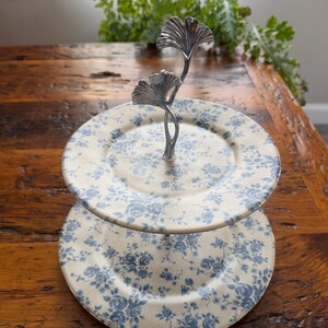 May include: Two-tiered serving stand with cream-colored plates adorned with blue floral patterns. The stand features a silver-toned metal handle with a leaf design. Ideal for displaying desserts or appetizers.