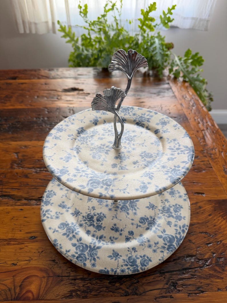 May include: Two-tiered serving stand with cream-colored plates adorned with blue floral patterns. The stand features a silver-toned metal handle with a leaf design. Ideal for displaying desserts or appetizers.