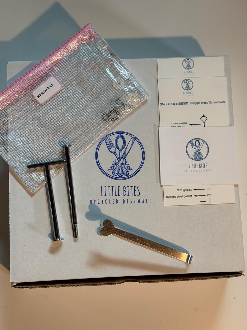 May include: A box containing upcycled dishware hardware. Includes a clear mesh bag labeled "HARDWARE", metal tools, and cards with instructions and the "LITTLE BITES UPCYCLED DISHWARE" logo. The box is white with blue text and graphics.