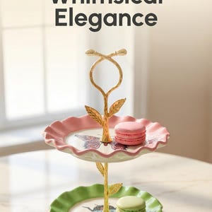 May include: Two-tiered serving stand with a gold-colored stem and floral accents. The top tier is pink, the bottom tier is green, and both have a scalloped edge. The stand holds two macarons. Text reads "*HANDCRAFTED*" and "A Touch of Whimsical Elegance."