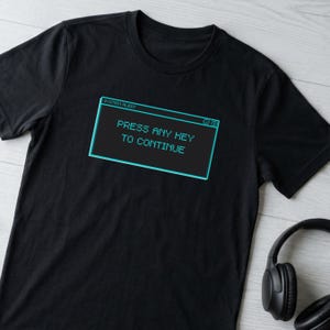 Puede incluir: Camiseta negra con un diseño de ventana de ordenador con bordes turquesa. El texto dice "SYSTEM ALERT" y "PRESS ANY KEY TO CONTINUE". La camiseta tiene cuello redondo y mangas cortas. Se ven unos auriculares negros.