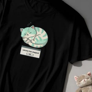 Könnte beinhalten: Schwarzes T-Shirt mit einem Cartoon-Kätzchen, das schläft, mit einem mintgrünen und weißen Streifenmuster. Der Text "cat.exe has stopped" wird in einer Fehlermeldung angezeigt, darunter das Wort "OK".