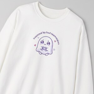 Könnte beinhalten: Weißes Sweatshirt mit Rundhalsausschnitt. Auf der Vorderseite befindet sich ein Cartoon-Geist, der einen Gamecontroller hält, mit dem Text "Haunted by bad teammates" in einem gebogenen Design. Der Geist ist lila mit rosa Herzen.