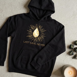 Könnte beinhalten: Schwarzer Kapuzenpullover mit einem goldenen Edelstein-Design und dem Text "LAST SAVE. NEVER." Die Grafik zeigt einen goldenen Edelstein mit strahlenförmigen Linien. Der Kapuzenpullover hat eine Vordertasche und eine Kapuze. Würfel und eine Kerze sind im Hintergrund.