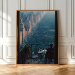 May include: A framed wall art print depicting a high-angle view from a balcony overlooking a city skyline and a large park. A person sits at a small metal table with a chair on the balcony, facing the urban landscape during the golden hour.