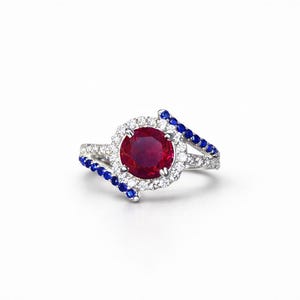May include: A silver ring with a large, round, red gemstone at the center, surrounded by smaller white gemstones. The ring has two curved bands, each lined with small blue gemstones. The ring is set against a white background.