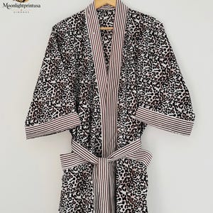 May include: A kimono robe featuring a black, white, and brown leopard print pattern. The garment includes a contrasting collar, sleeve cuffs, and a waist tie, all decorated with a vertical brown and white pinstripe design on a light background.