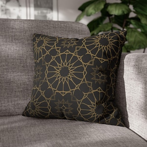 May include: Square throw pillow with a black background and a gold geometric star pattern. The pillow is placed on a gray sofa, offering a decorative accent for home decor.
