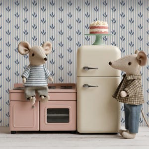 May include: Two plush mouse dolls in a miniature kitchen setting. One mouse wears a blue and white striped shirt and grey shorts, sitting on a pink toy stove. The other mouse wears a brown plaid jacket and blue trousers. A small cake sits on a cream refrigerator.
