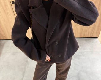 May include: A dark brown double-breasted blazer with structured shoulders and notched lapels, featuring two dark buttons on the front and two smaller buttons on each cuff, paired with matching dark brown trousers and light tan slip-on shoes.