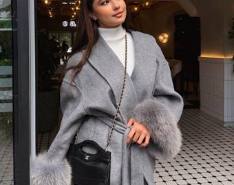 May include: A gray wrap coat featuring long sleeves with thick gray fur cuffs, a tie waist, and a white mock neck top underneath. A small black handbag with a gold chain strap is worn across the body, paired with solid black pants.