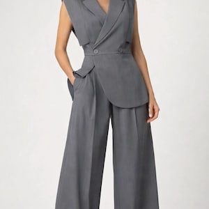 K&ouml;nnte beinhalten: Graues &auml;rmelloses Blazer- und Hosen-Set mit weitem Bein. Der Blazer hat ein doppelreihiges Design mit Wickel-Front und Schulterpolstern. Die Hose ist hoch tailliert mit weitem, flie&szlig;endem Bein. Das Outfit ist aus einem einfarbigen grauen Stoff gefertigt.