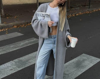 May include: A long, open-front grey knit cardigan with wide sleeves, paired with a white cropped tank top, light-wash blue denim jeans, and pointed-toe shoes. The outfit is accessorized with gold hoop earrings and clear-framed glasses.