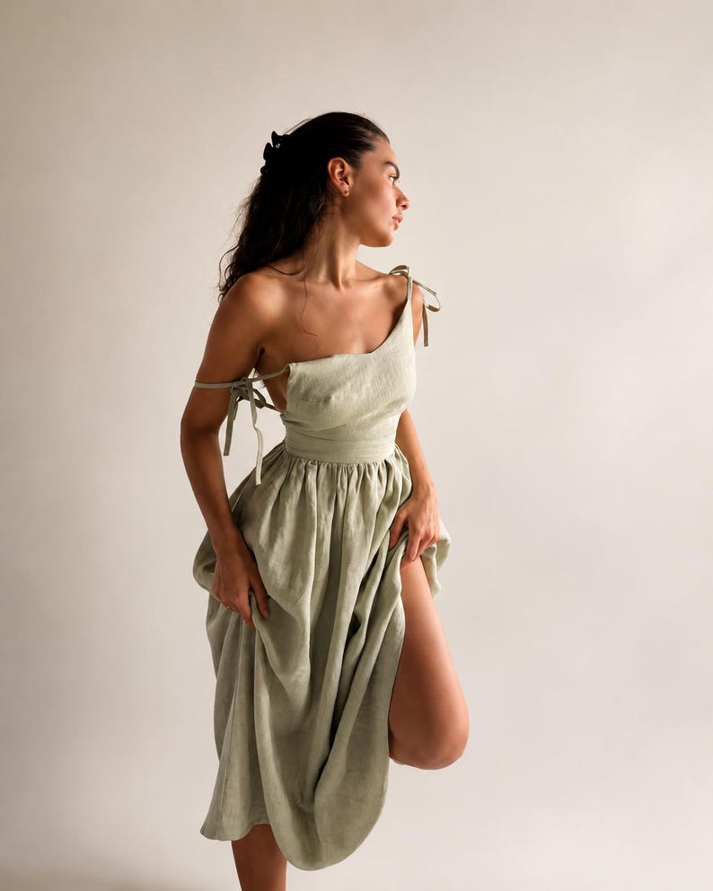 May include: A light green linen dress with a one-shoulder design and tie straps. The dress features a fitted bodice and a flowing skirt with a high slit. The model is wearing the dress in a studio setting.