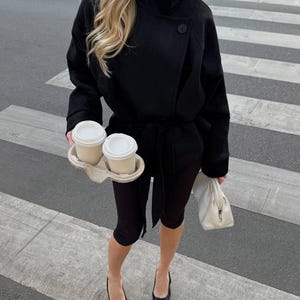 May include: A black belted jacket with a high collar and long sleeves, paired with black leggings and black pointed-toe heels. The outfit is accessorized with a white clutch bag and a cardboard carrier holding two white disposable coffee cups.