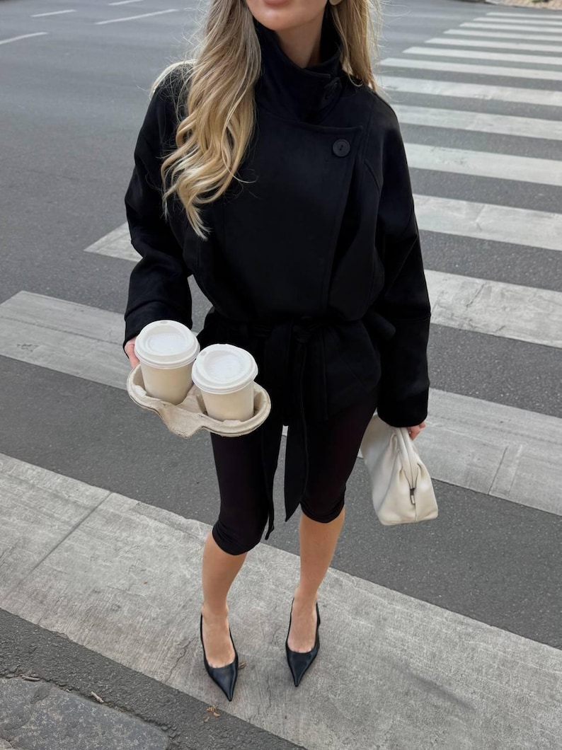 May include: A black belted jacket with a high collar and long sleeves, paired with black leggings and black pointed-toe heels. The outfit is accessorized with a white clutch bag and a cardboard carrier holding two white disposable coffee cups.