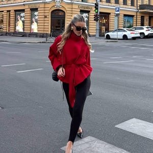 May include: A bright red long-sleeved jacket with a high collar and a matching fabric tie belt at the waist. The jacket is paired with black stirrup leggings and black pointed-toe heels. A small black quilted shoulder bag is carried on the side.