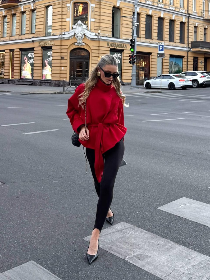 May include: A bright red long-sleeved jacket with a high collar and a matching fabric tie belt at the waist. The jacket is paired with black stirrup leggings and black pointed-toe heels. A small black quilted shoulder bag is carried on the side.