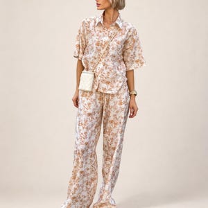 Puede incluir: Un conjunto a juego con estampado floral, compuesto por una camisa de manga corta y pantalones de pernera ancha. El conjunto presenta un fondo crema con un estampado floral marr&oacute;n. Se lleva un peque&ntilde;o bolso blanco acolchado con una cadena dorada.