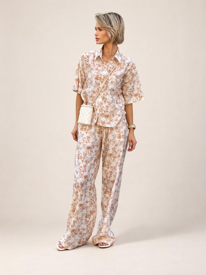 Puede incluir: Un conjunto a juego con estampado floral, compuesto por una camisa de manga corta y pantalones de pernera ancha. El conjunto presenta un fondo crema con un estampado floral marr&oacute;n. Se lleva un peque&ntilde;o bolso blanco acolchado con una cadena dorada.