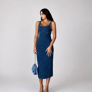May include: A dark blue denim midi dress featuring a square neckline, thick shoulder straps, and vertical seam detailing along the bodice and skirt. The dress is paired with a matching blue denim quilted handbag with a silver chain strap and blue mules.