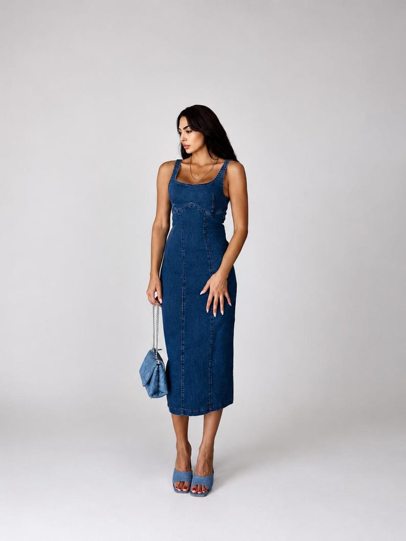 May include: A dark blue denim midi dress featuring a square neckline, thick shoulder straps, and vertical seam detailing along the bodice and skirt. The dress is paired with a matching blue denim quilted handbag with a silver chain strap and blue mules.