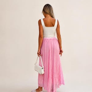 May include: A white sleeveless tank top with a square neckline paired with a long, flowing, semi-sheer pink skirt with a crinkled texture. The outfit includes a small white shoulder bag and white flat shoes, viewed from the back.