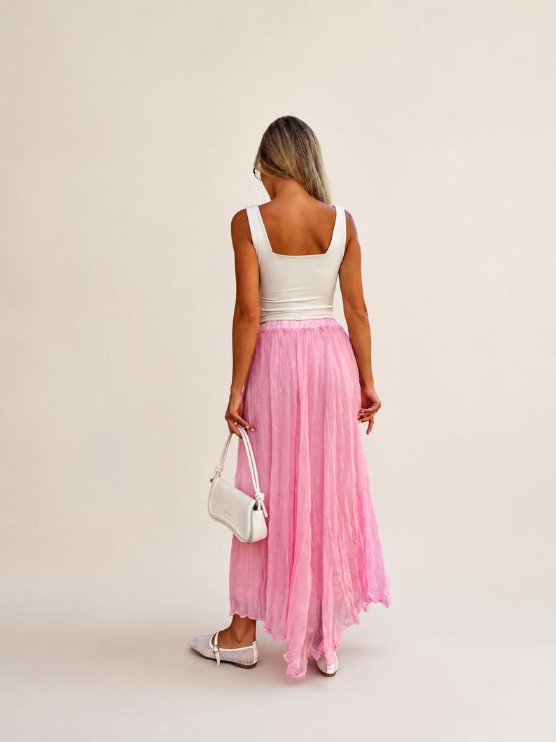 May include: A white sleeveless tank top with a square neckline paired with a long, flowing, semi-sheer pink skirt with a crinkled texture. The outfit includes a small white shoulder bag and white flat shoes, viewed from the back.