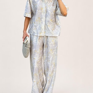 Puede incluir: Conjunto de dos piezas azul claro con un estampado floral en amarillo y blanco. El conjunto incluye una camisa de manga corta y pantalones de pernera ancha. La camisa tiene un bolsillo en el pecho. Los pantalones son largos.