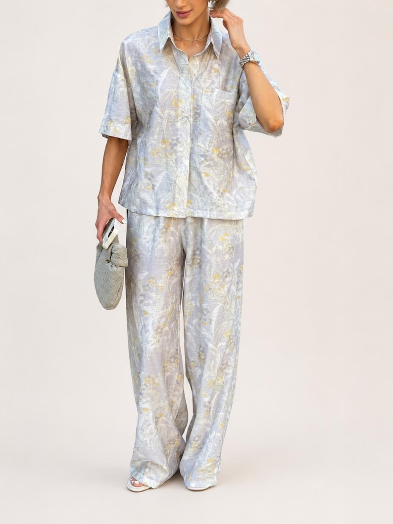 Puede incluir: Conjunto de dos piezas azul claro con un estampado floral en amarillo y blanco. El conjunto incluye una camisa de manga corta y pantalones de pernera ancha. La camisa tiene un bolsillo en el pecho. Los pantalones son largos.
