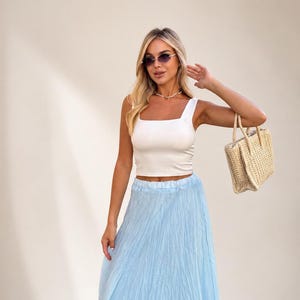 May include: A white sleeveless crop top with a square neckline paired with a light blue crinkled maxi skirt. The outfit is accessorized with a woven straw handbag, a delicate necklace, and dark-tinted sunglasses with thin gold-toned frames.